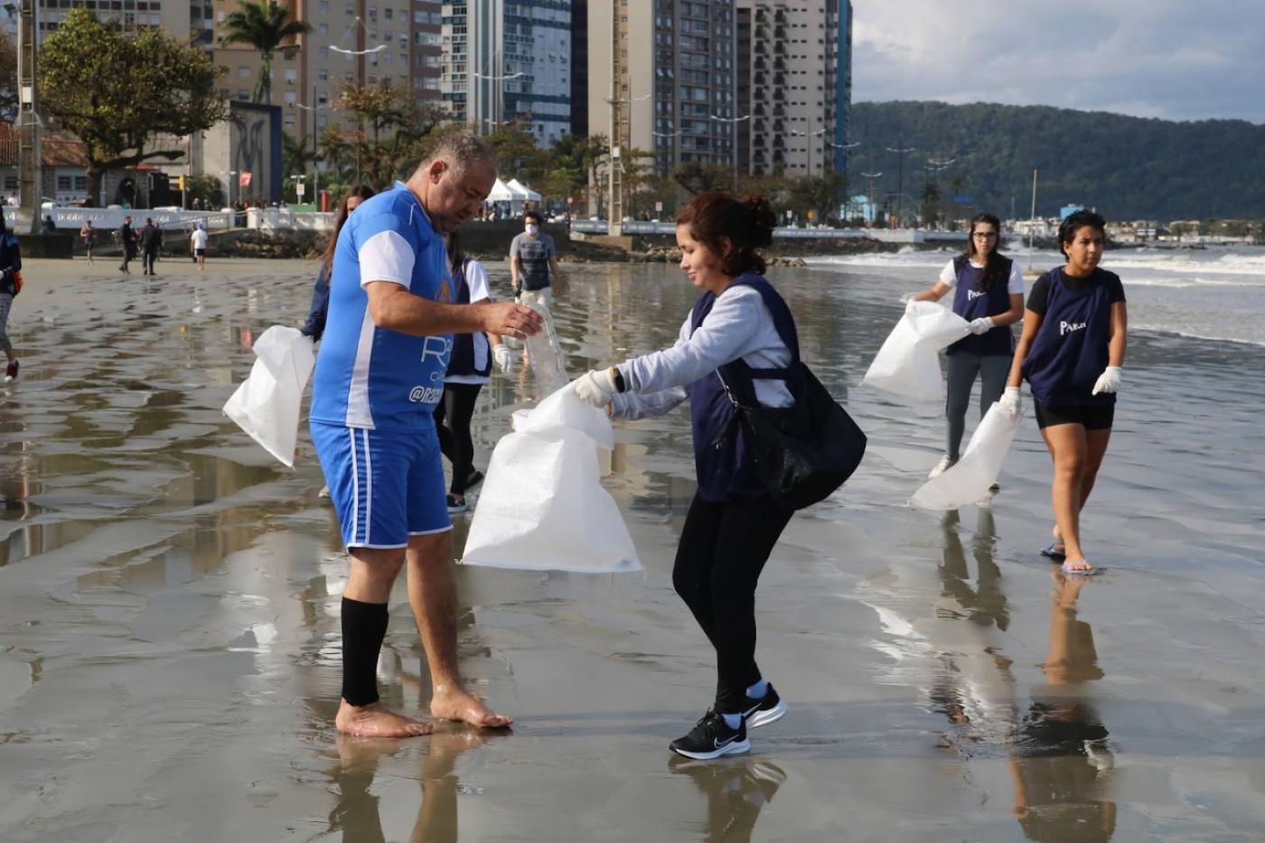 jovens estão na beira do mar segurando sacos de lixo onde depositam material coletado. #paratodosverem