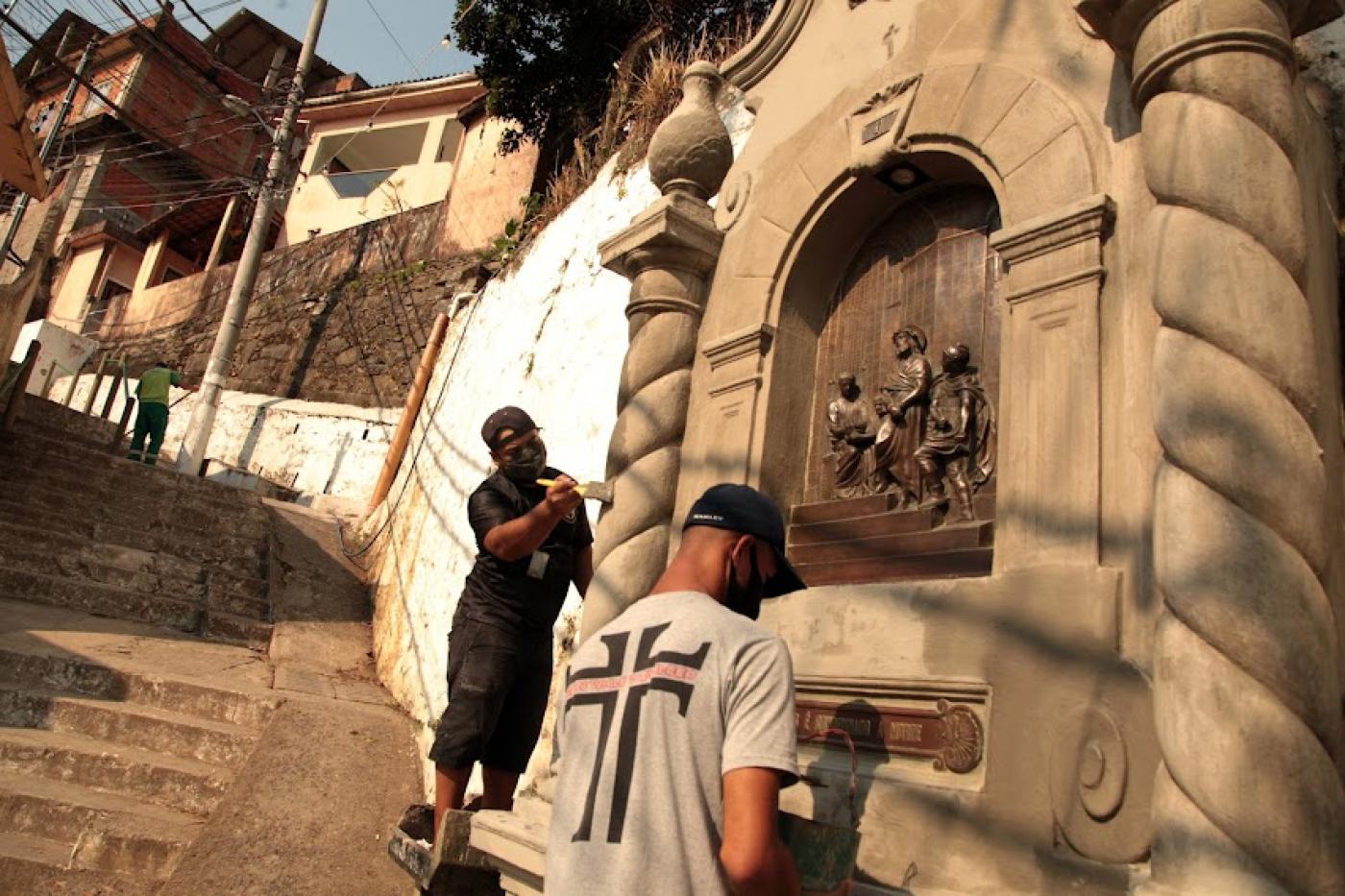 homem limpam nicho em muro. Em primeiro plano, um rapaz está de costas. Na camisa está estampado um crucifixo, numa ligação com o tema da reportagem, a via crucis. #paratodosverem