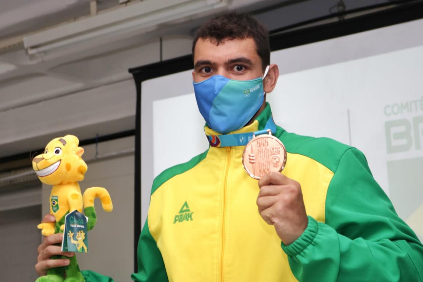 O atleta segura o boneco do mascote da competição na mão direita e a medalha na esquerda. Ele está de uniforme e máscara. #paratodosverem