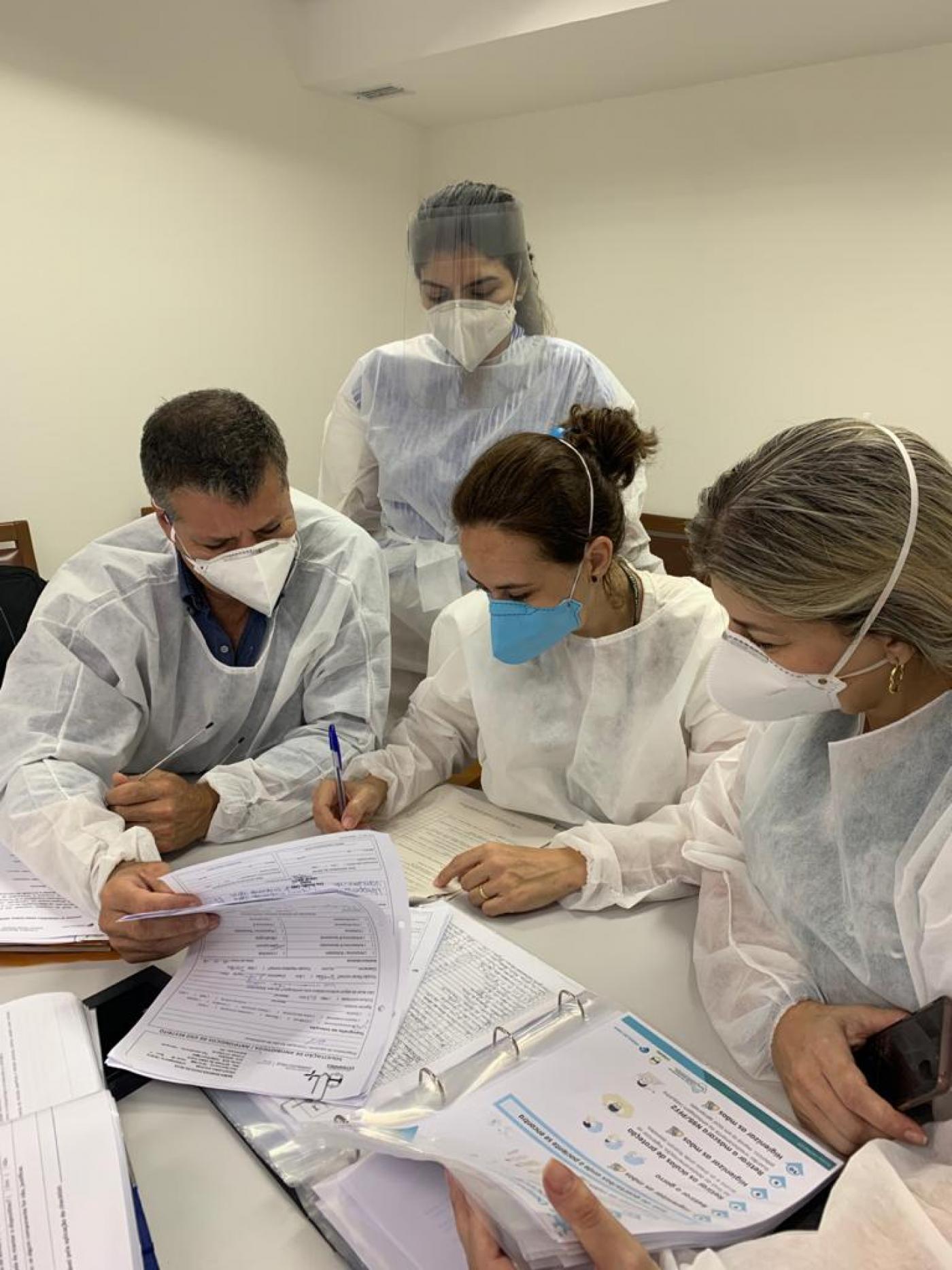 agentes de saúde paramentados estão mexendo em papeis com anotações em cima de uma mesa. #Paratodosverem