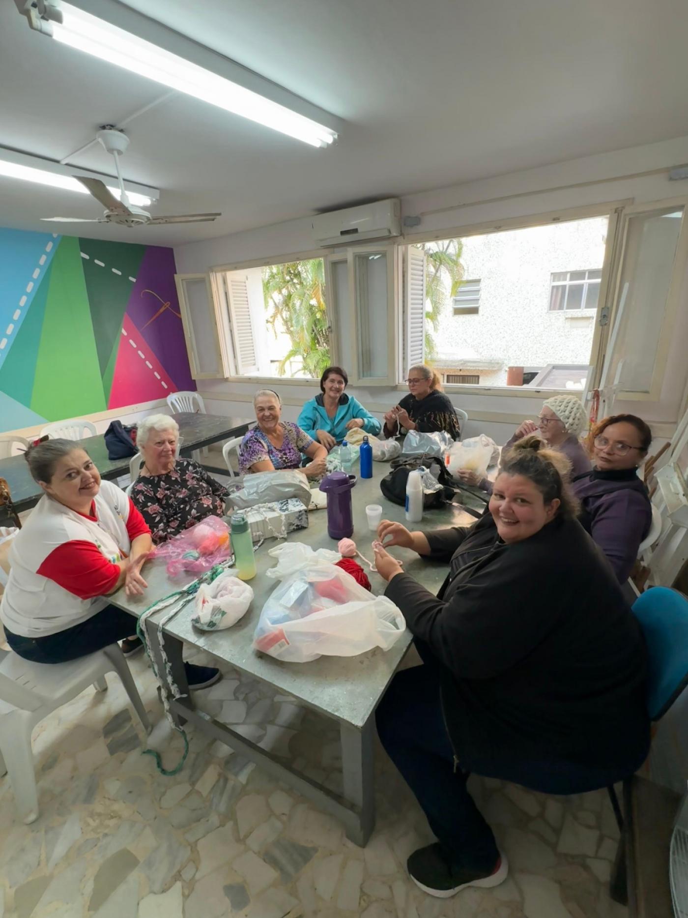 mulheres estão reunidas em torno de mesa fazendo artesanato. Algumas olham para a foto e sorriem. #paratodosverem 