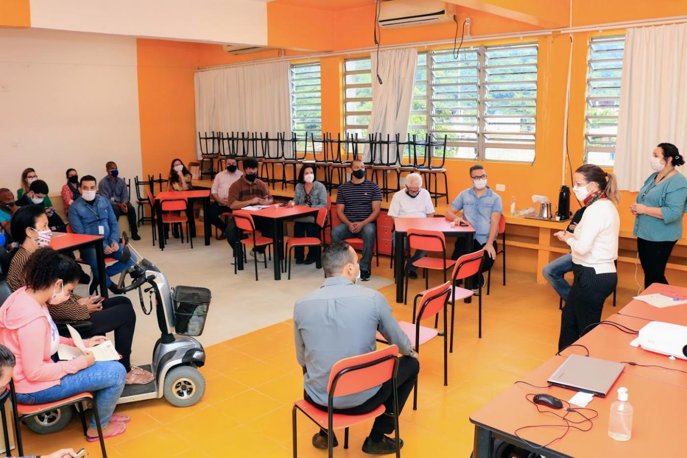 Sala de aula com cadeiras dispostas em círculo. Pessoas estão sentadas distantes umas das outras. #paratodosverem