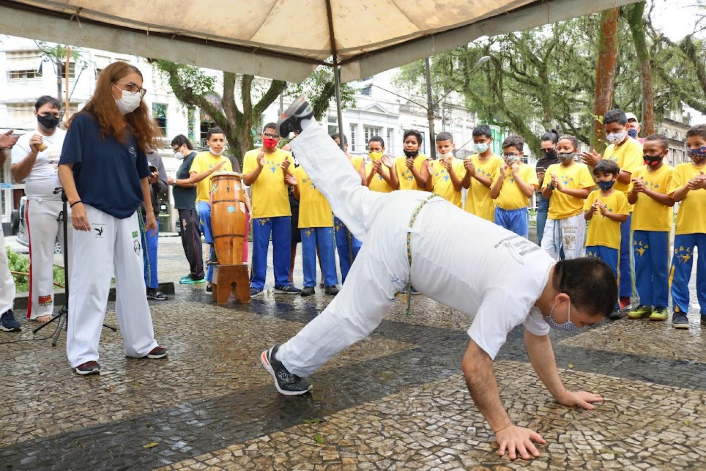 homem está jogando capoeira com as duas mãos e um dos pés no chão. A outra perna está no ar. Ele está de costas. O público aplaude ao fundo. #paratodosverem