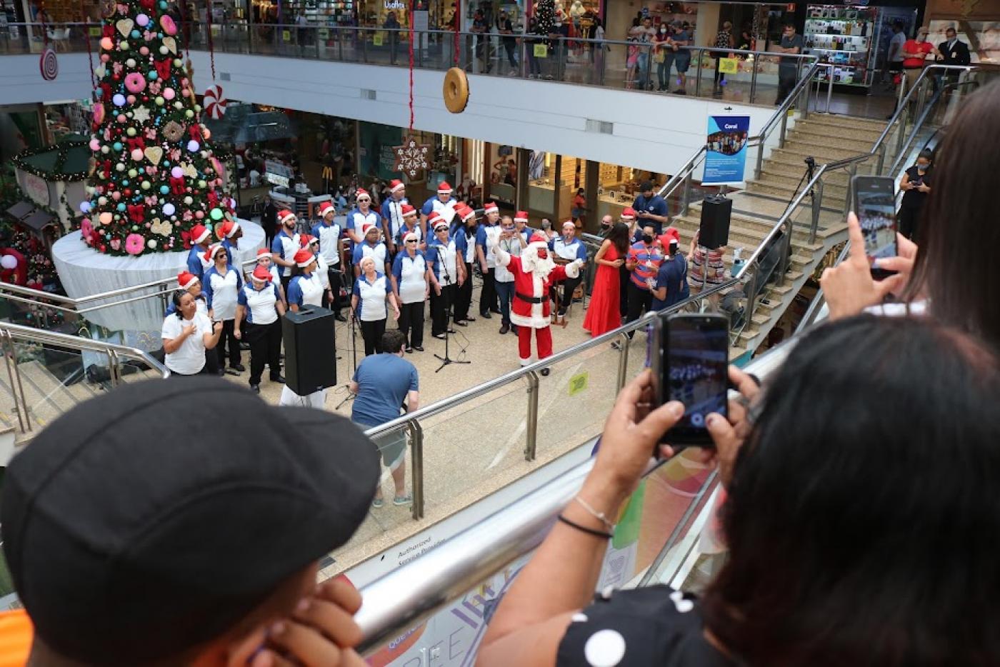 integrantes de coral estão em formação de apresentação com uma grande árvore de natal ao fundo. Em primeiro plano, o público tira fotos da apresentação com celulares. #paratodosverem