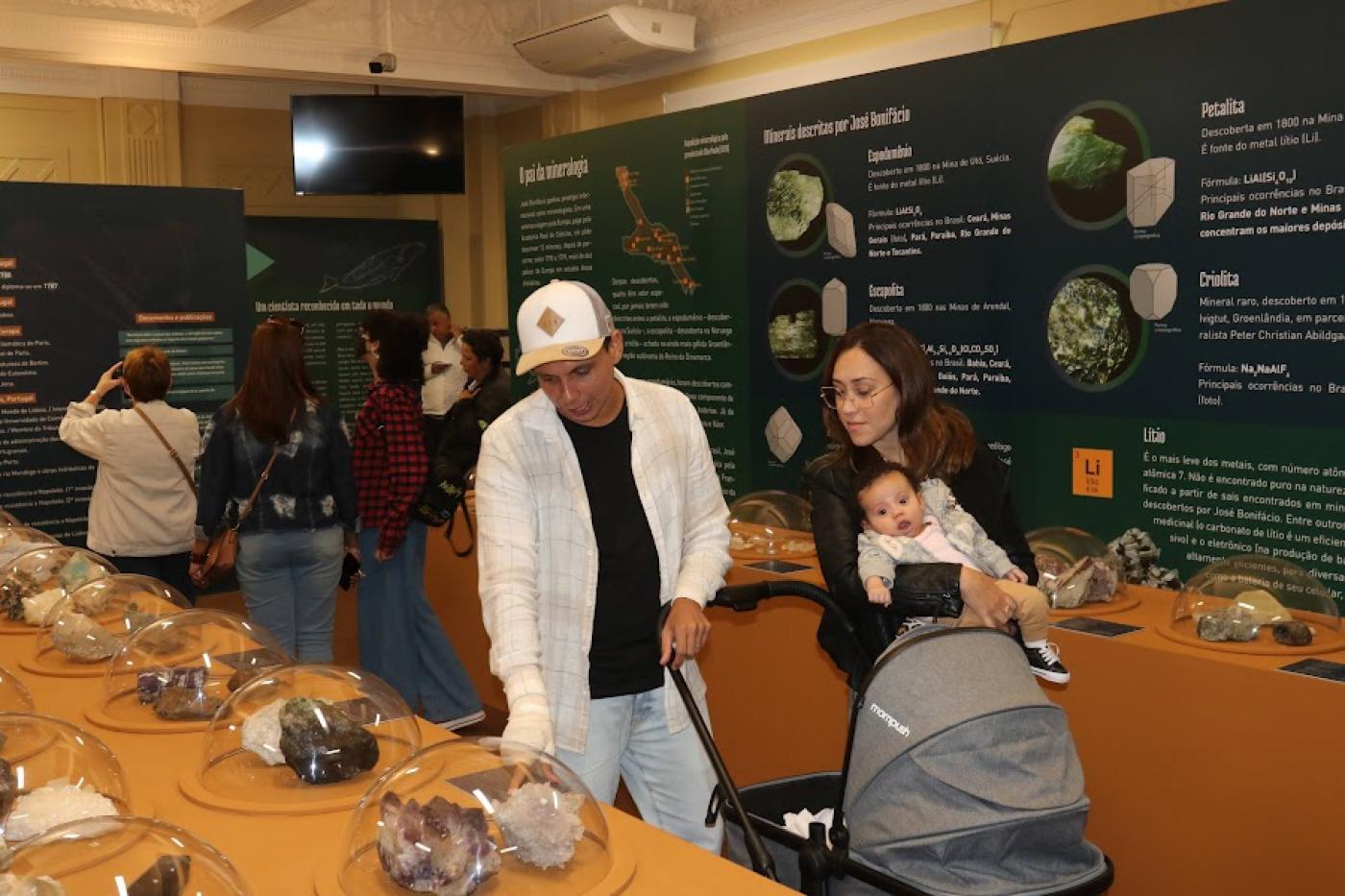 sala de exposição com várias cúpulas transparentes cobrindo pedaços de pedras. Um casal com um bebê está olhando a exposição. Há outras pessoas ao fundo. #paratodosverem