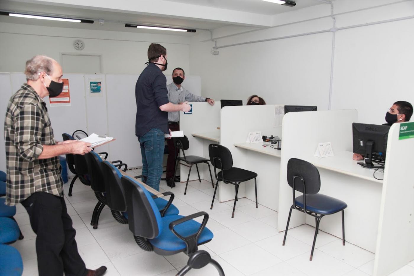 Dois homens, um deles segurando um papel, estão dentro de uma sala com três funcionários. Todos usam máscaras. #Paratodosverem