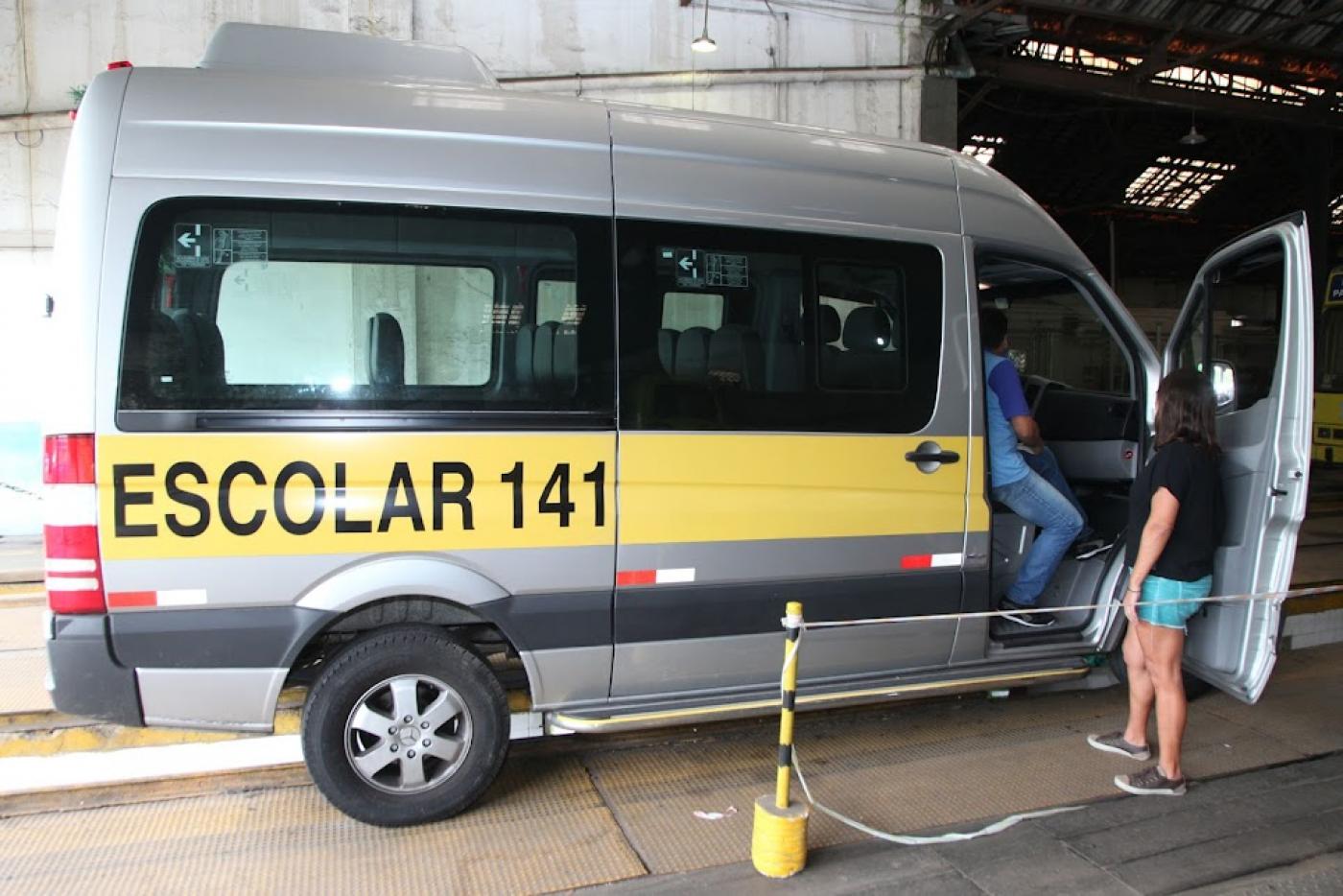 van escolar está parada em baia. Há uma pessoa sentada no banco do passageiro olhando o interior do veículo. Outra, está encostada na porta observando. #paratodosverem