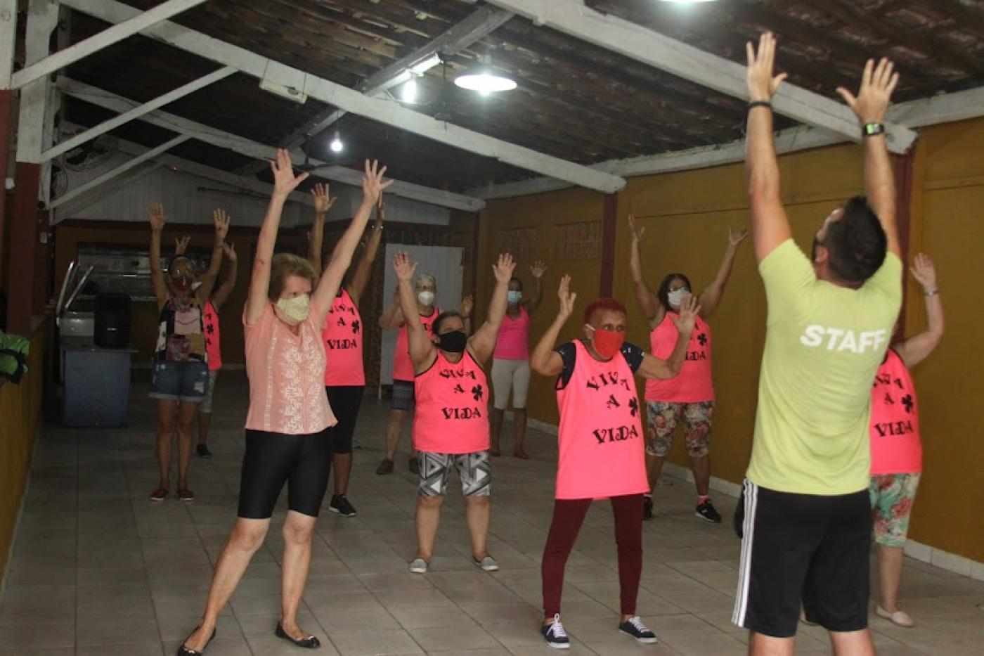 alunas estão em pé, todas com os braços para o alto, fazendo igual ao professor que está à frente delas e de costas para a foto. #paratodosverem