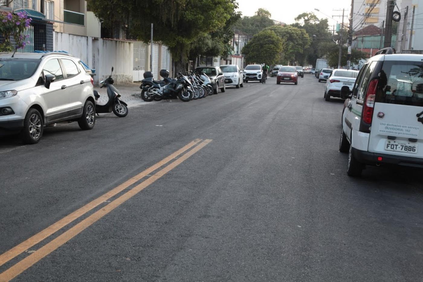 trecho de rua com asfalto novo e pintura de solo. Carros estão estacionados dos dois lados. #paratodosverem