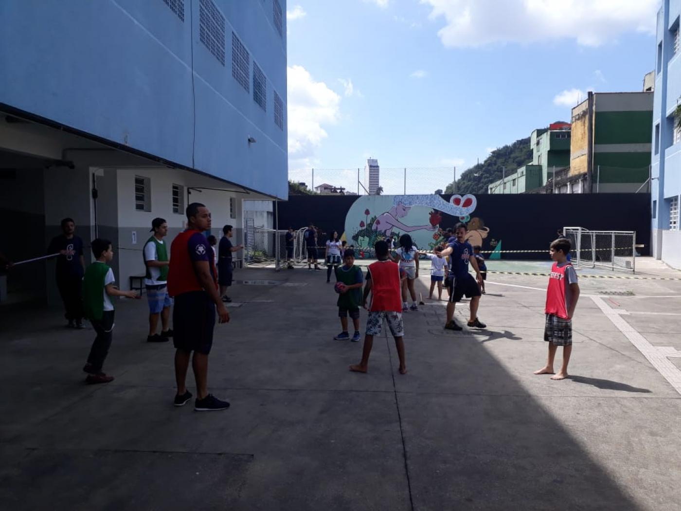 Crianças brincam na quadra da escola. Elas jogam bola com as mãos e usam coletes para diferenciar os times #Pracegover