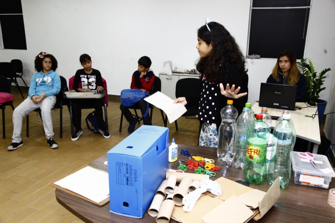 Crianças estão em uma sala. Uma delas está em pé ao lado de vários produtos recicláveis, como garrafas PET, rolos e papel e outros objetos. #Pracegover