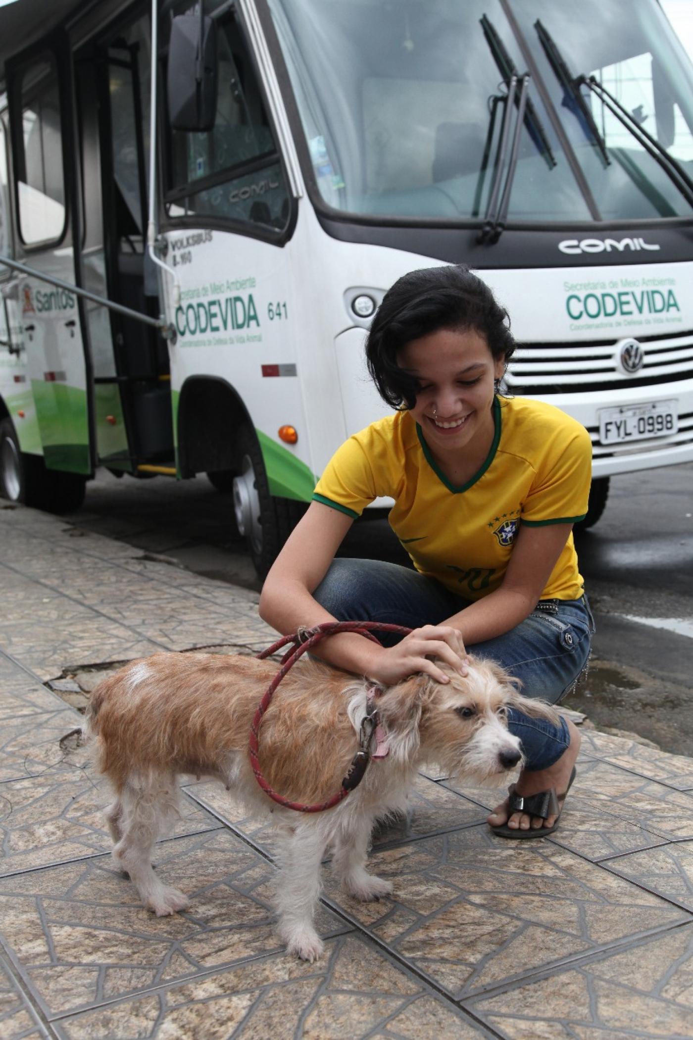 Mulher está agachada e passa a mão na cabeça de um cachorro. Ao fundo está o castramóvel. #Pracegover