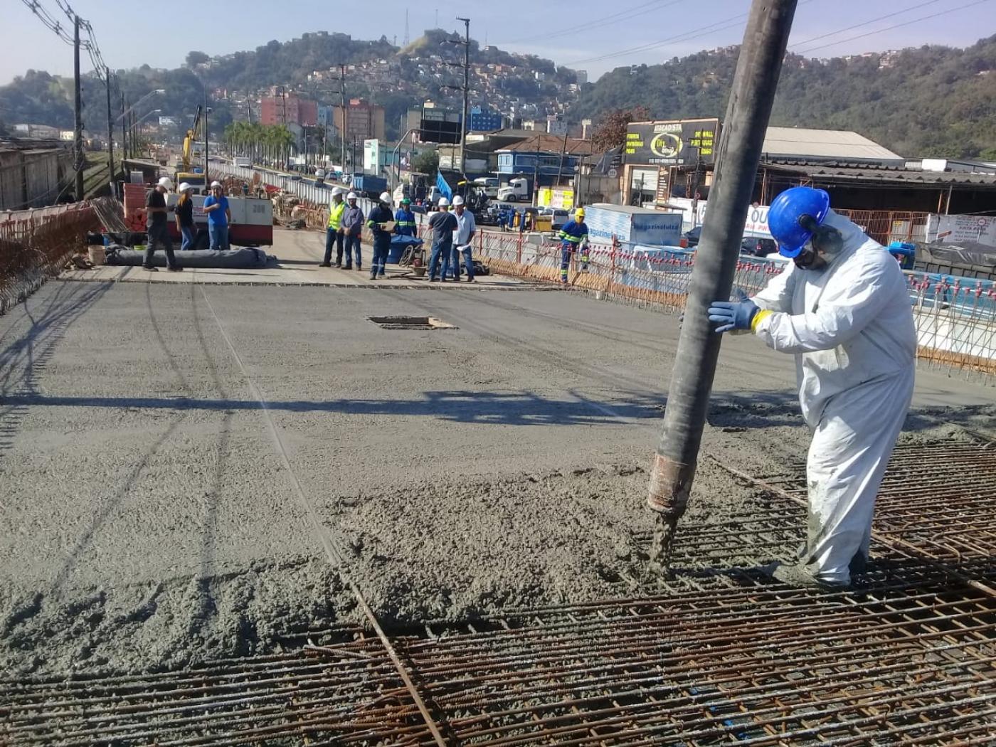 Homem com uniforme branco e capacete azul manuseia mangueira que despeja concreto sobre pista de viaduto