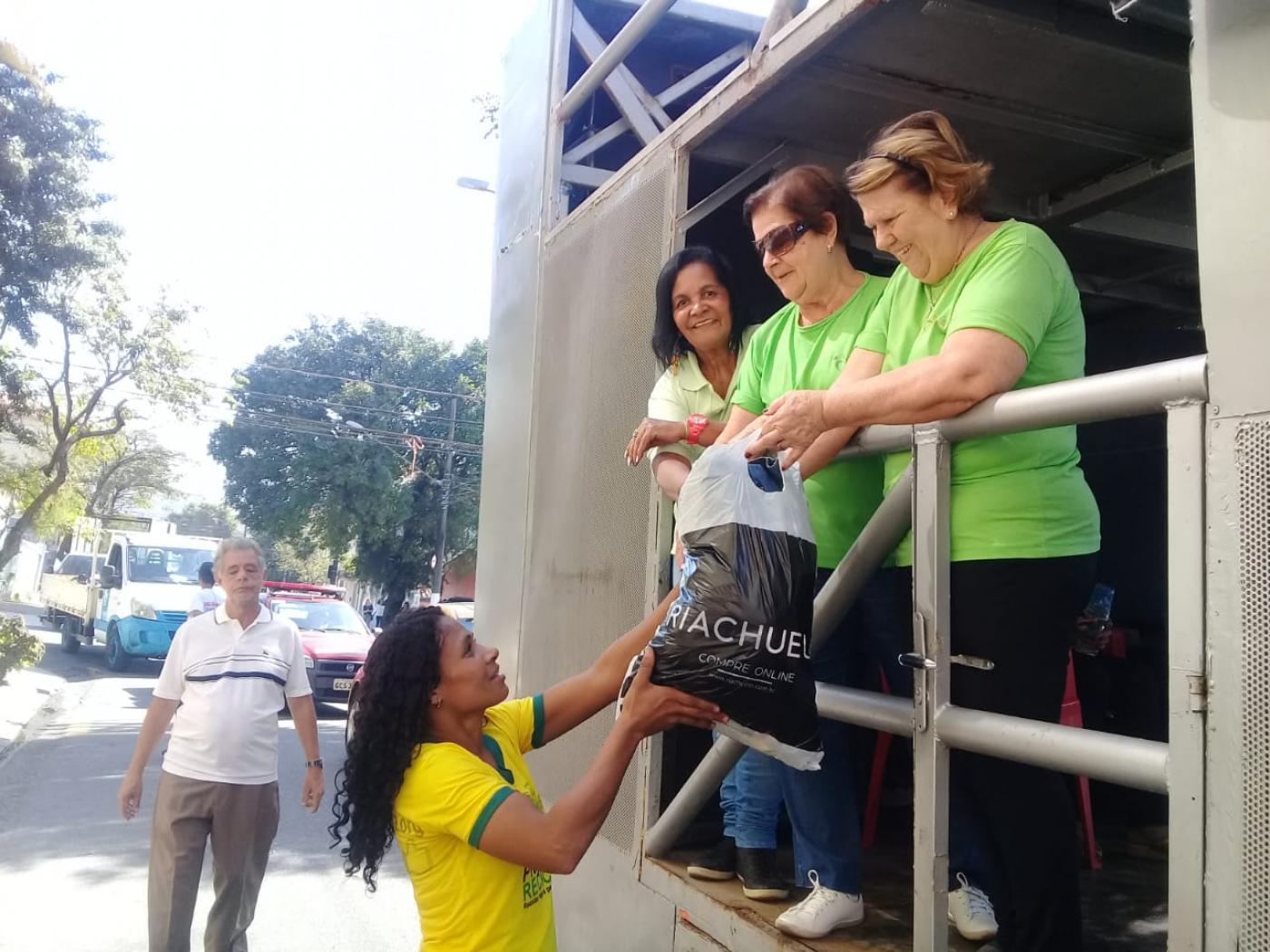 Com camisa da seleção brasileira de futebol, mulher entrega sacola com roupas para três mulheres de verde sobre caminhão