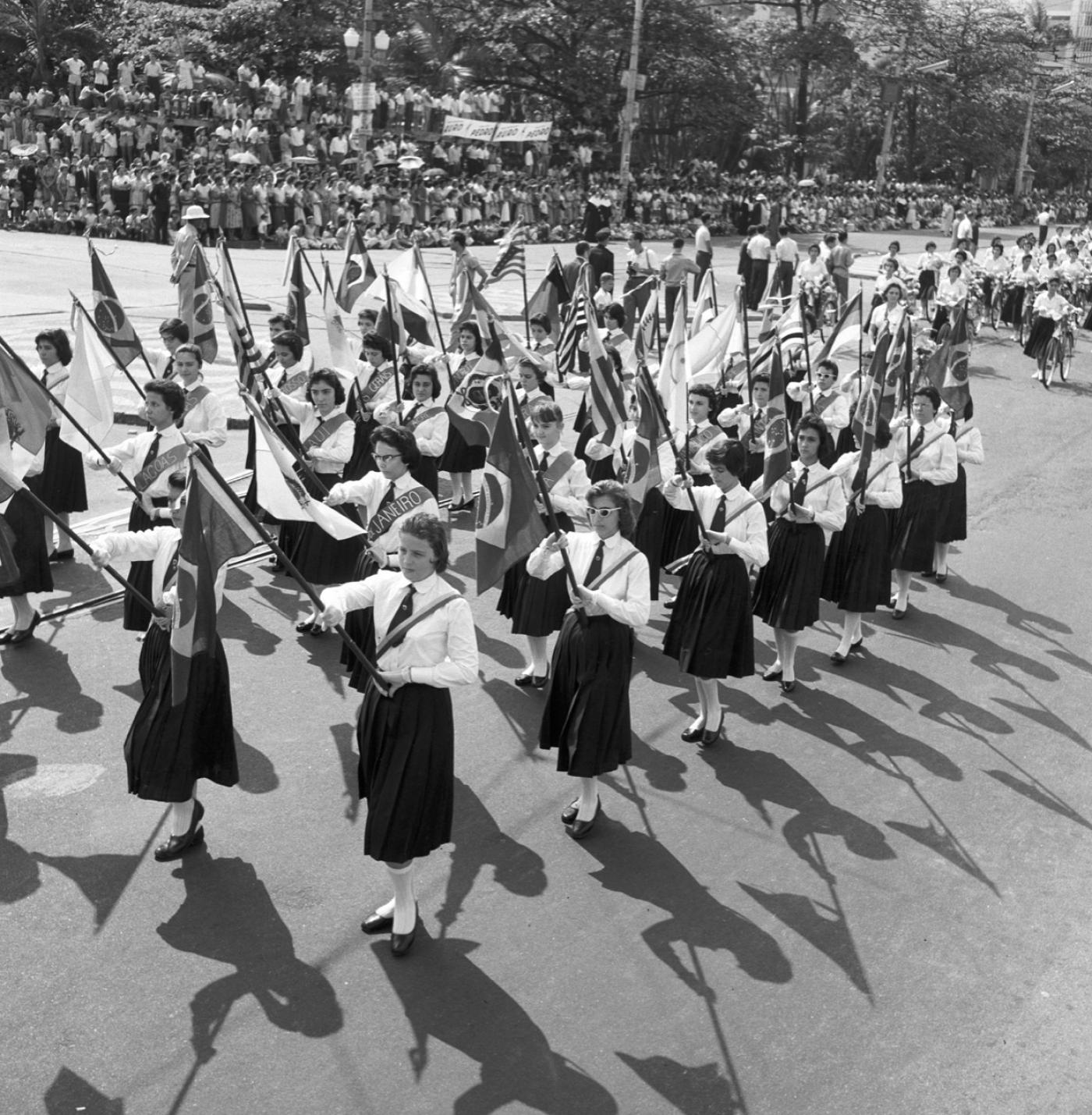 Antigo desfile em Santos. Mulheres vestidas com saias e usando gravatas seguram bandeiras. #Pracegover