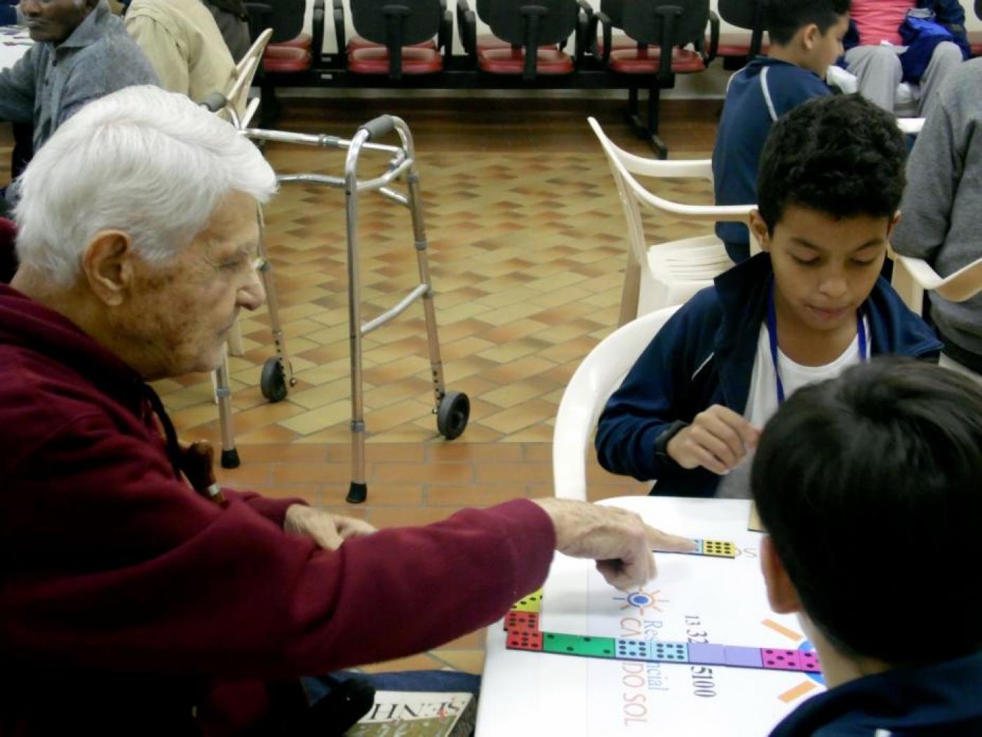 idoso joga dominó sentado em mesa com dois estudantes #paratodosverem