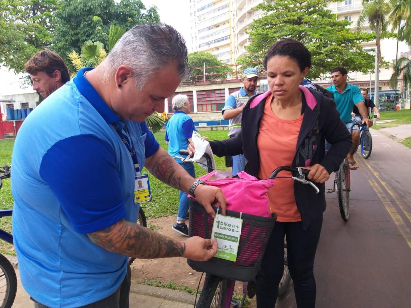 Agente da CET coloca placa em biclicleta. Uma mulher está desmontada da bike aguardando a afixação. Atrás há movimento de bicicletas e outros agentes. #Pracegover