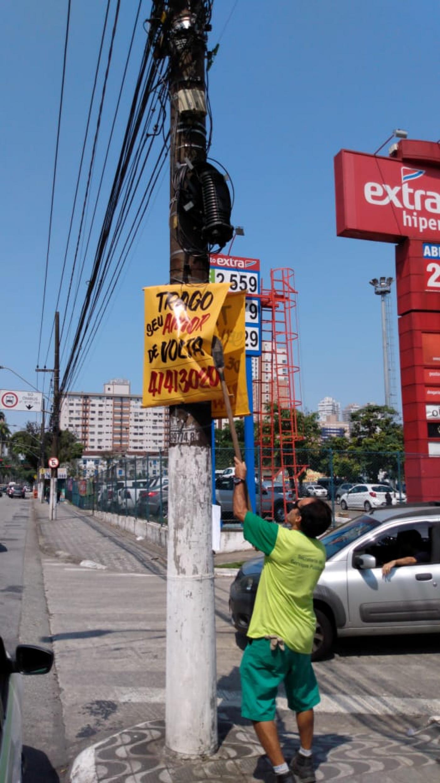 Homem remove banner afixado em poste. Ele usa uma vara para alcançar a propaganda. #Pracegover