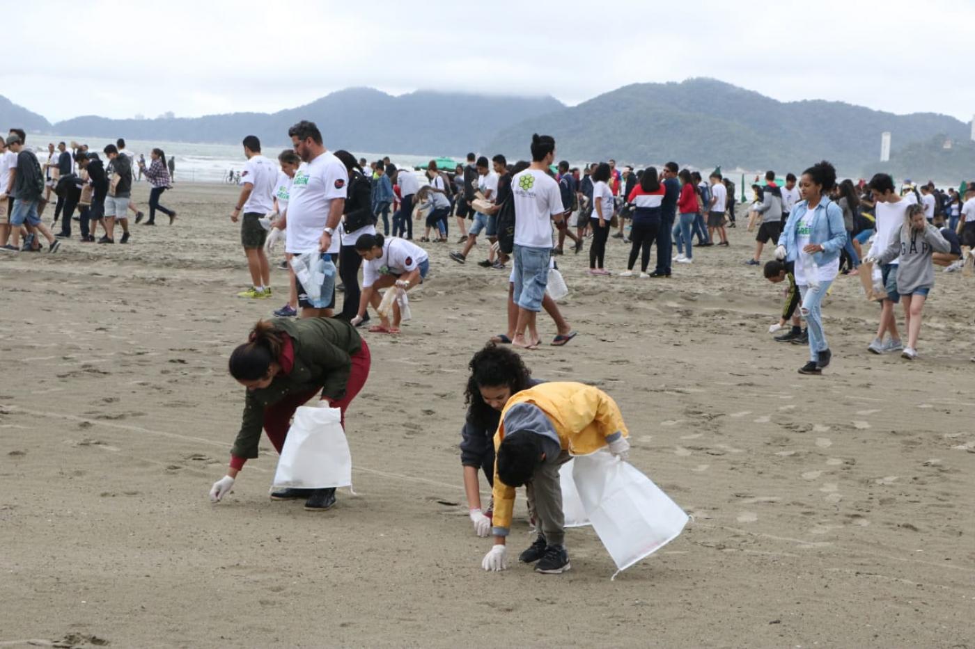 Três pessoas agachadas recolhem lixo da areia da praia, com cerca de 100 pessoas ao fundo vom sacolas plásticas nas mãos
