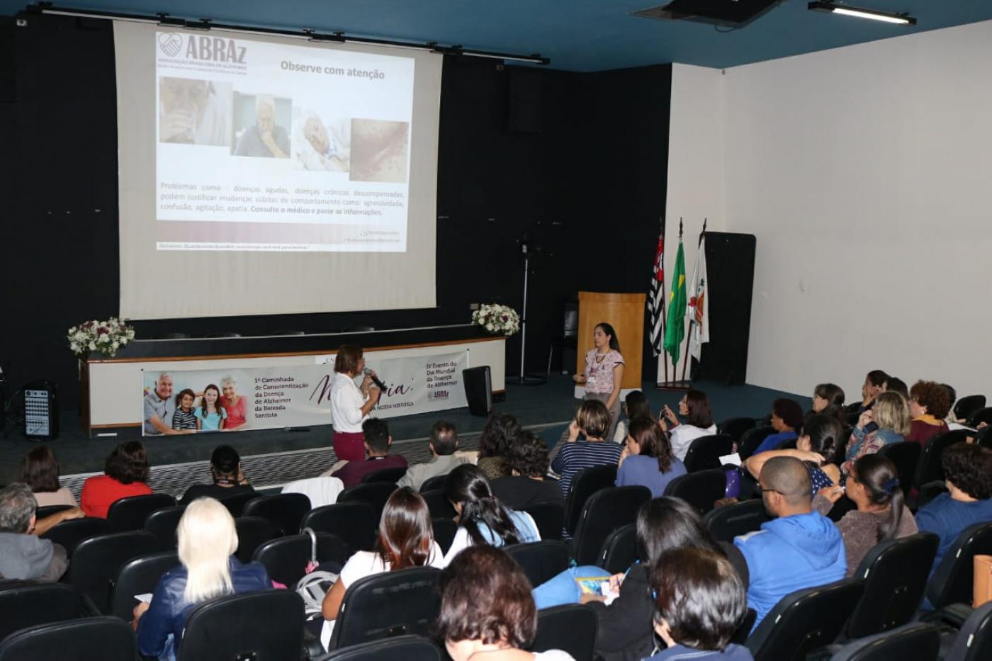 Cerca de 30 pessoas sentadas em auditório assitem a mulher com microfone falando à frente de projeção