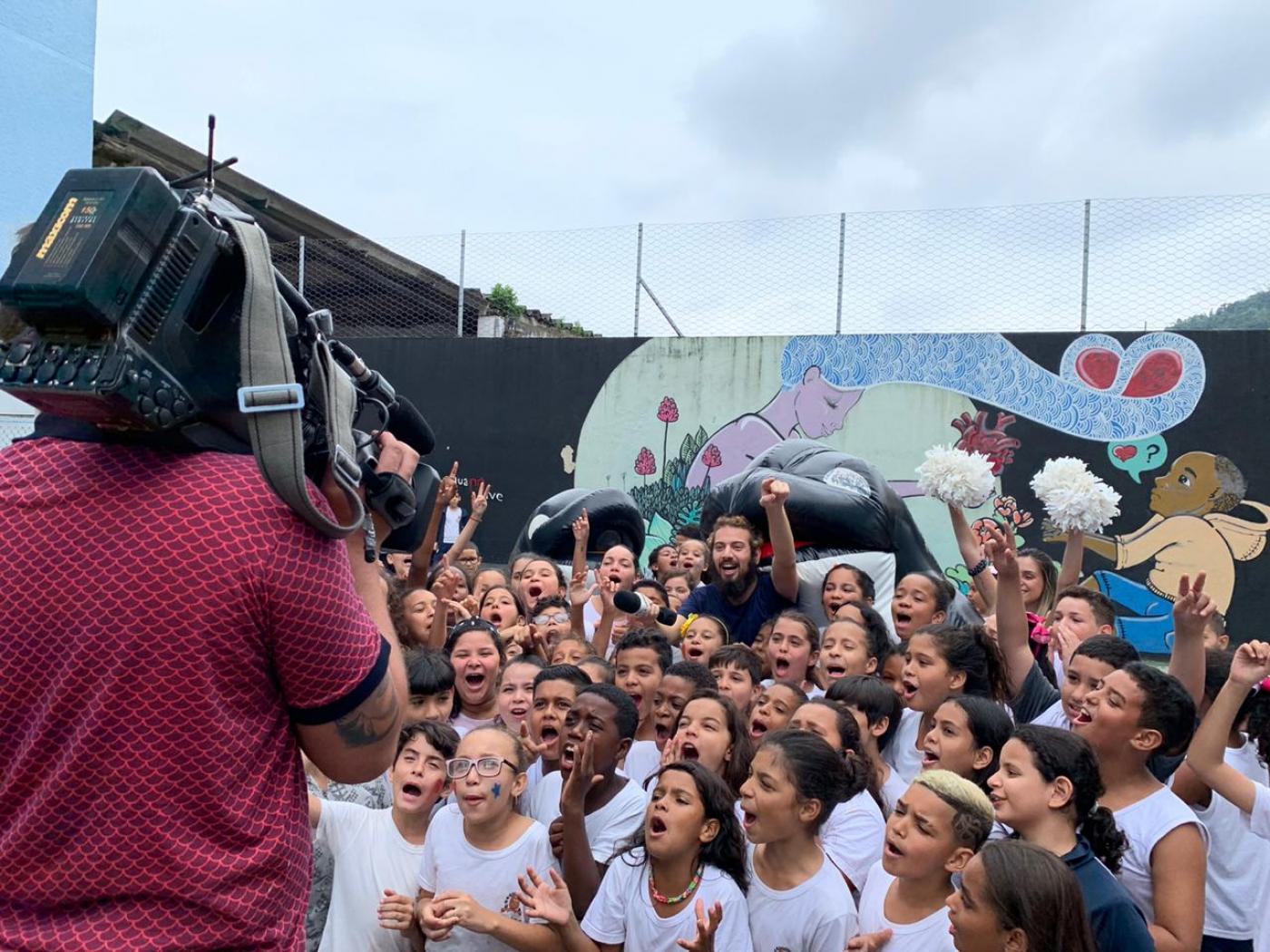 Cinegrafista grava cerca de 50 crianças com uniforme escolar em frito de guerra com jornalista ao fundo