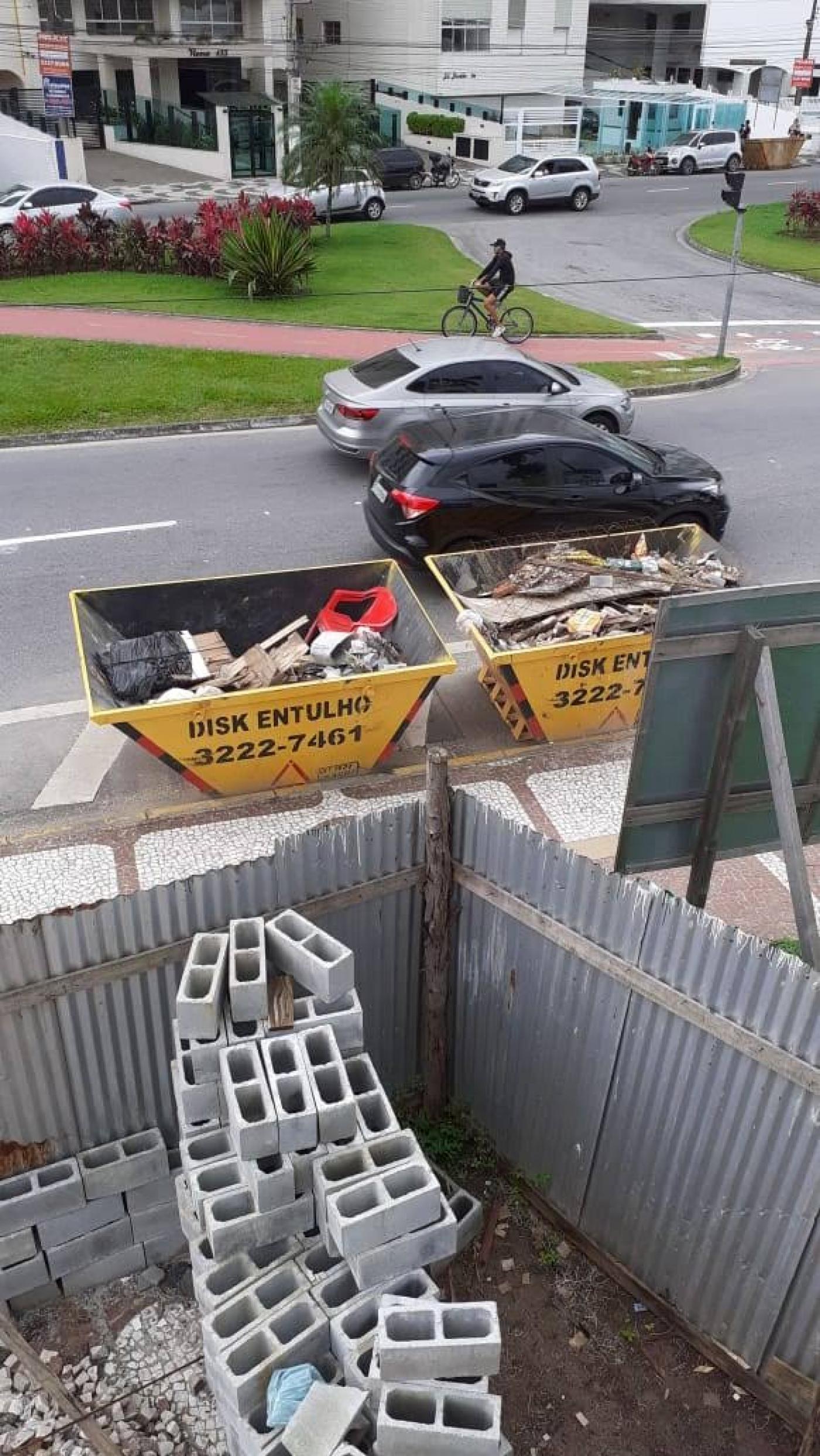 Tijolos vazados para obra em primeiro plano. Ao fundo, do lado de fora da construção, há duas caçambas com material depositado. #Pracegover