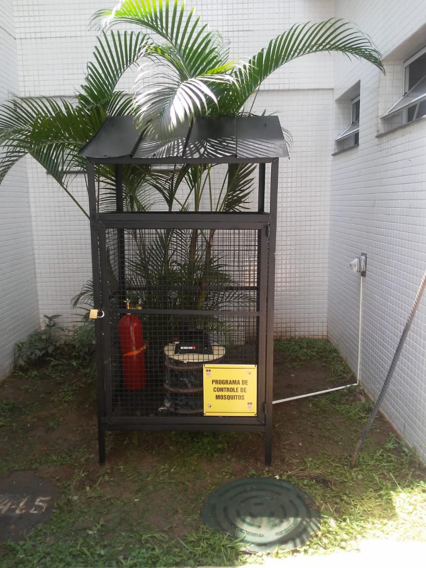 Contadora automática de mosquitos instalada em uma pequena área com vegetação. O material está dentro de uma cabine de grades. #Pracegover
