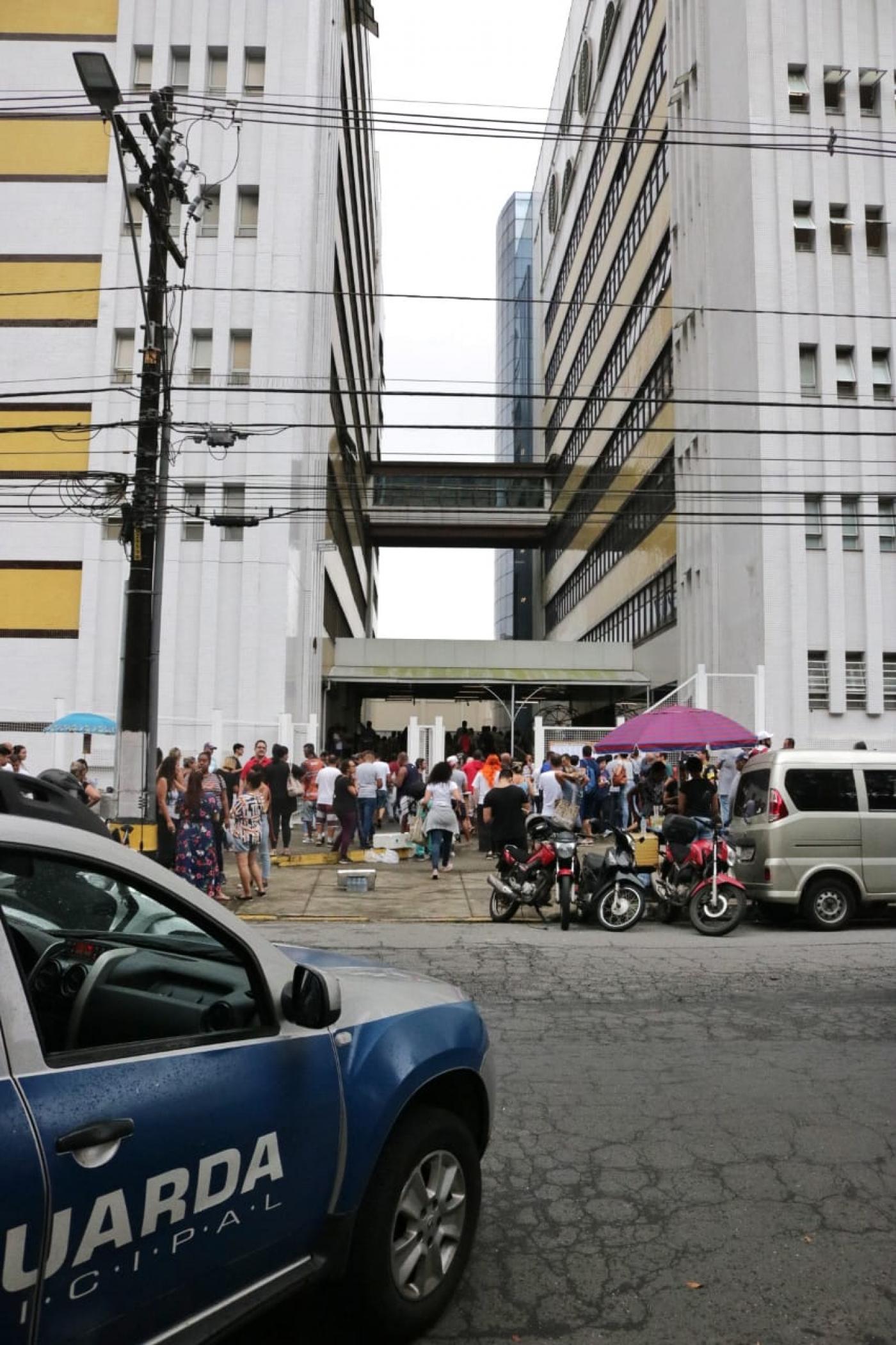 #pracegover Com carro da Guarda Municipal em primeiro plano, foto mostra dezenas de pessoas entrando em prédio de universidade