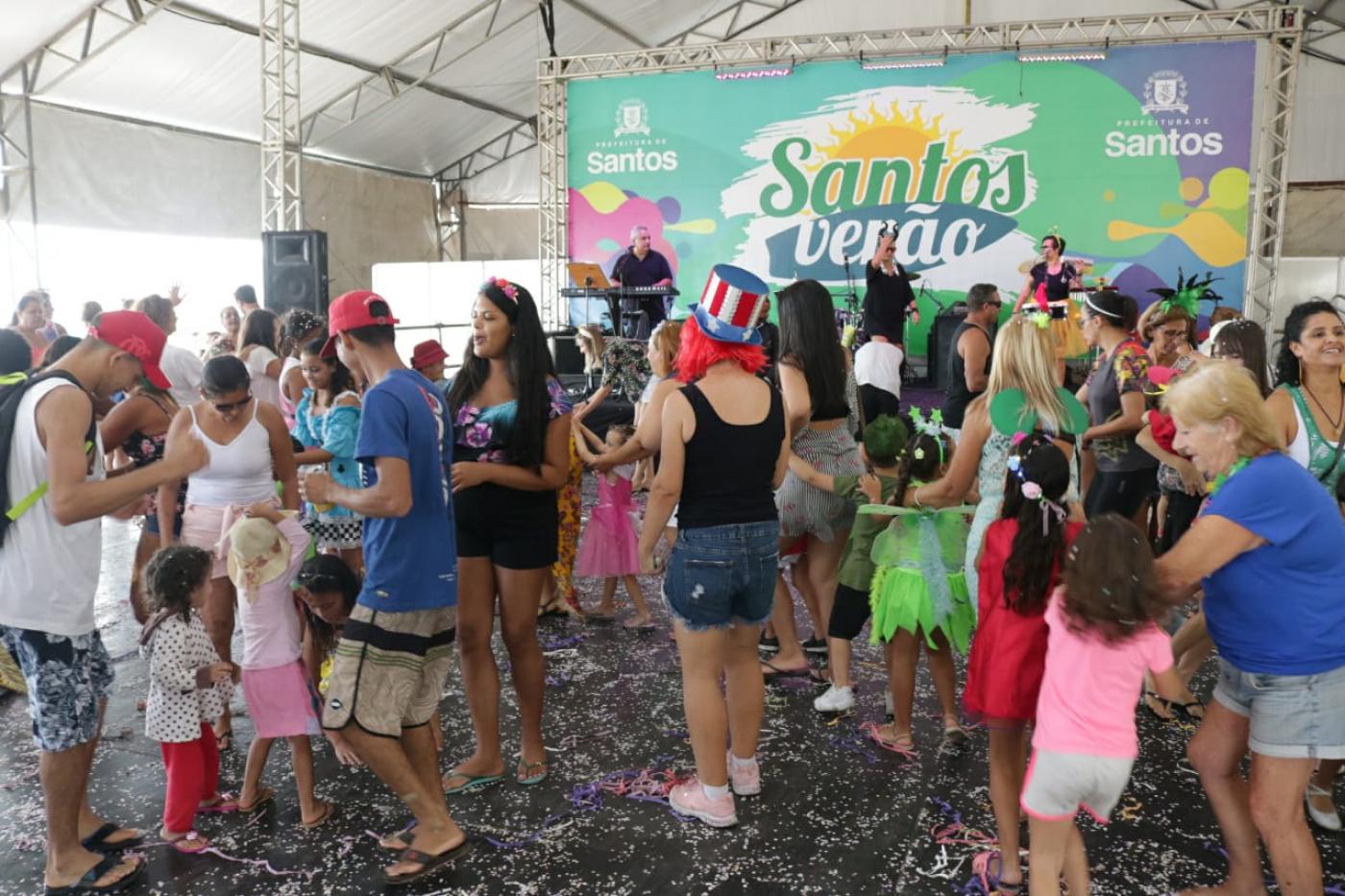 #pracegover Sobre chão repleto de confetes, dezenas de adultos e crianças fantasiados brincam no interior de tenda