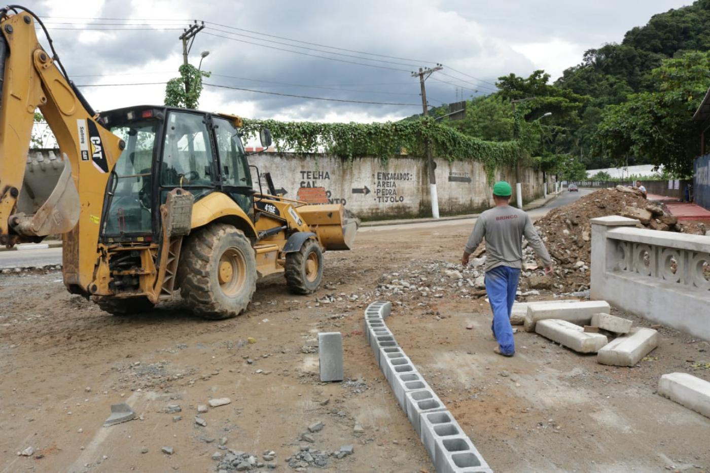 #pracegover homem com capacete caminha ao lado de escavadeira em terreno com materiais de construção junto à avenida