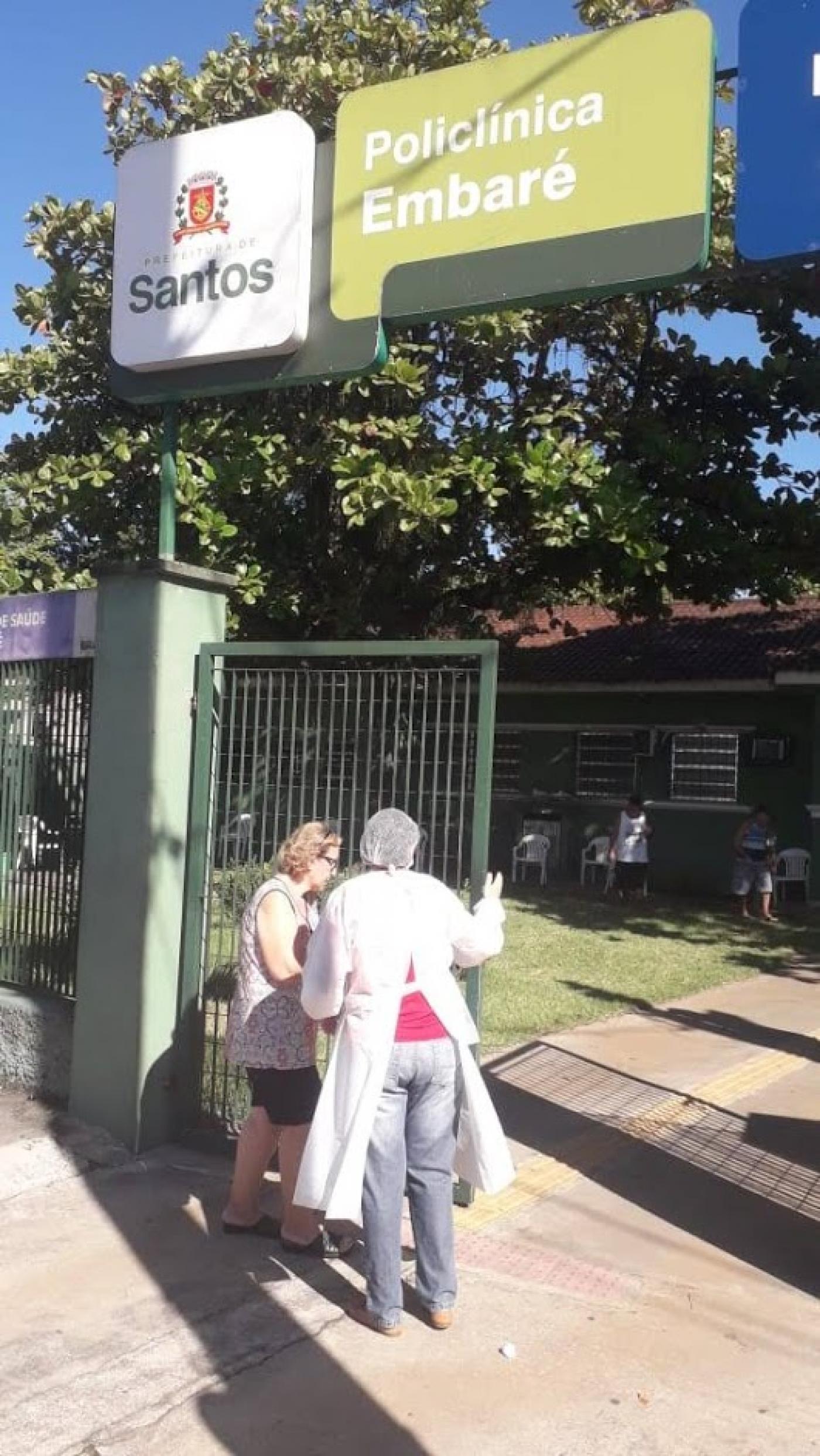 Fachada de policlínica com uma mulher entrando e outra, com avental, touca e luvas a orientando. #Paratodosverem