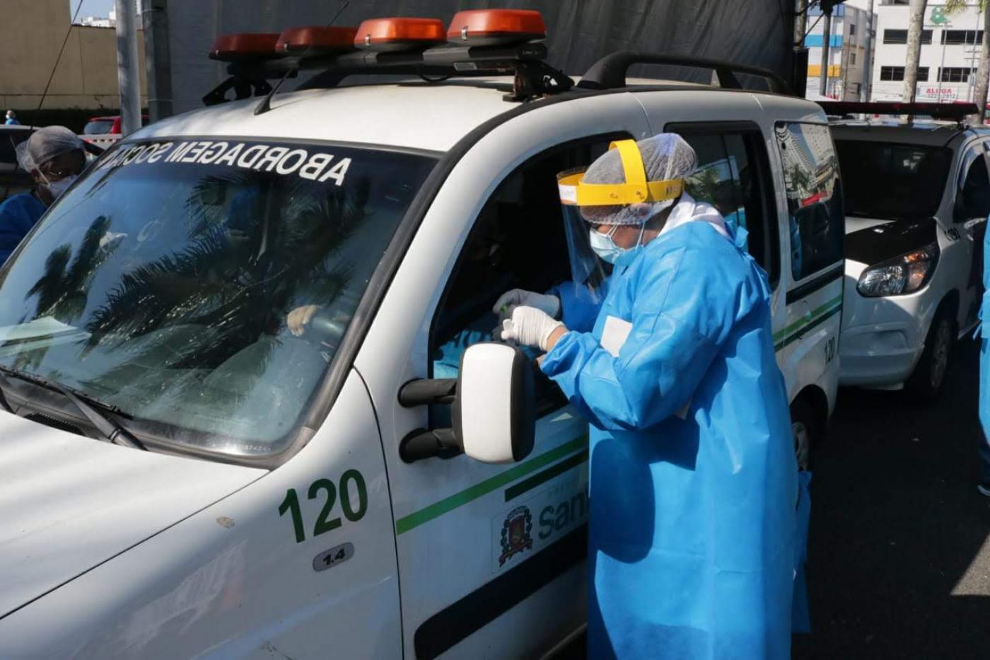 #pracegover com capa azul e proteção facial, enfermeira aplica teste em motorista pela janela do veículo sob tenda