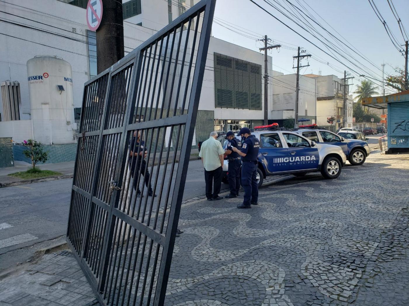 Portão de alumínio encostado em um posto e ao lado guardas e viatura da guarda. #paratodosverem