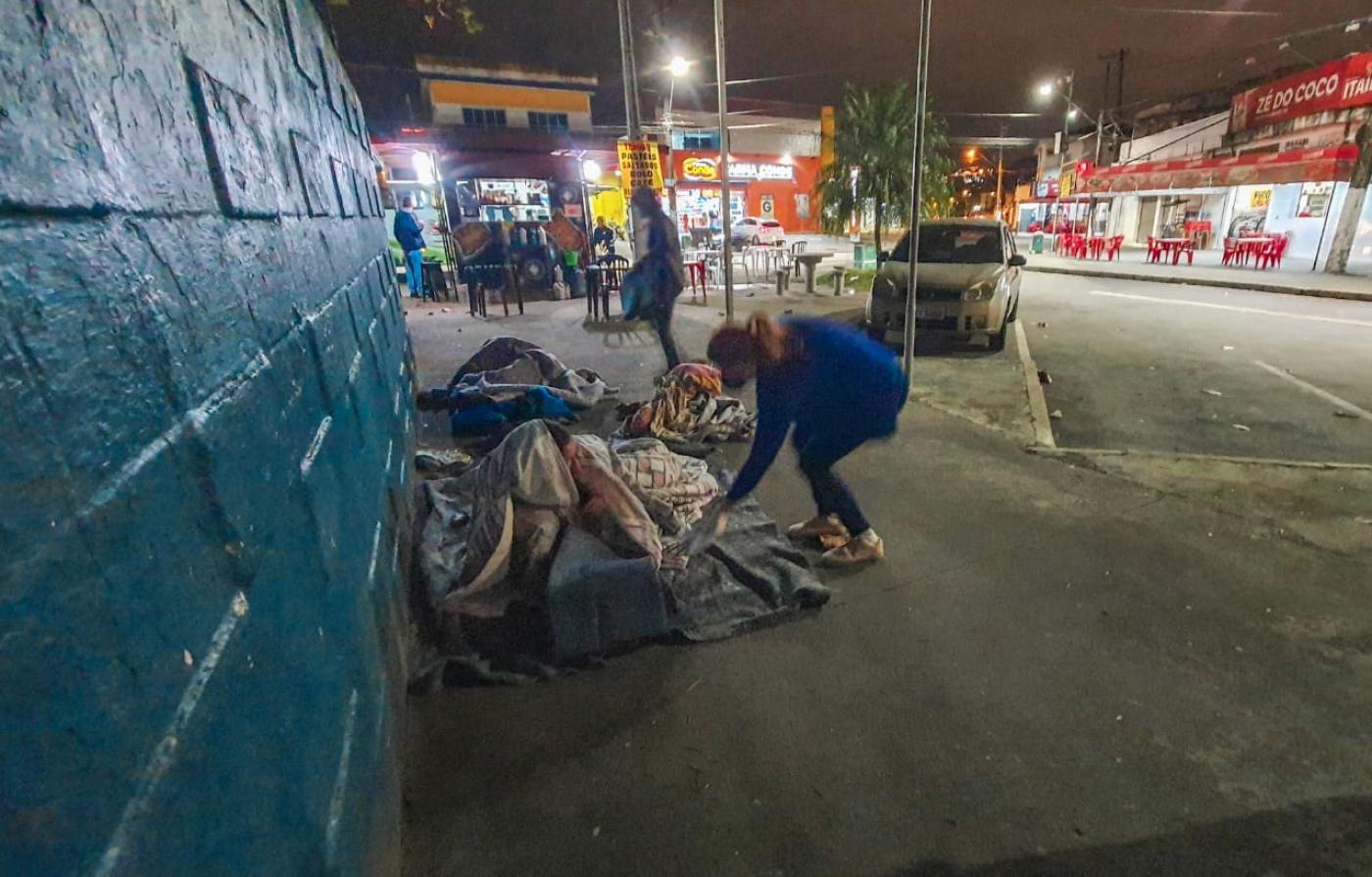 #pracegover Sobre a calçada, mulher entraga cobertor a homem deitado