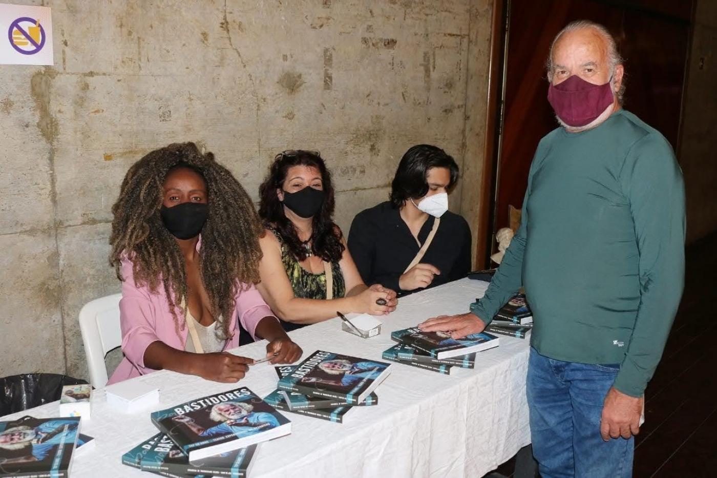 homem encosta em livros sobre a mesa observado por 3 mulheres #paratodosverem