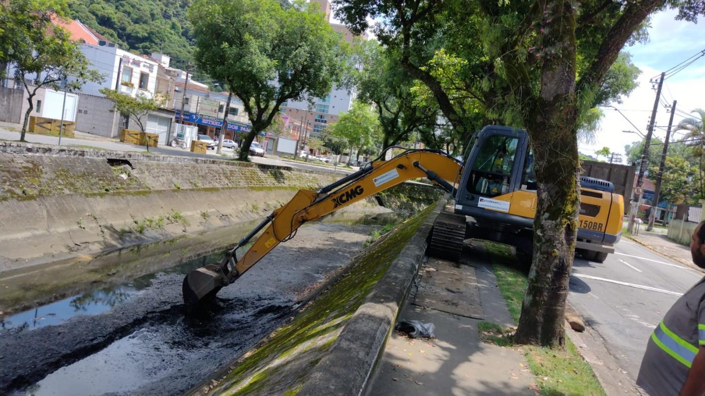 escavadeira parada em calçada recolhe lama de fundo de canal. A escavadeira está na calçada entre duas árvores. #paratodosverem