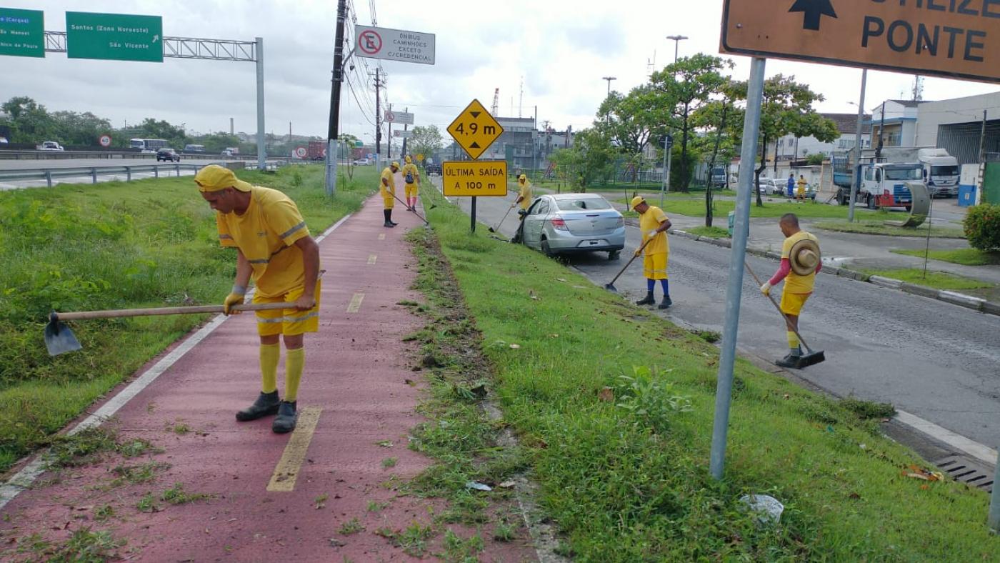 pessoas uniformizadas de amarelo cortando mato em área junto à rodovia #paratodosverem