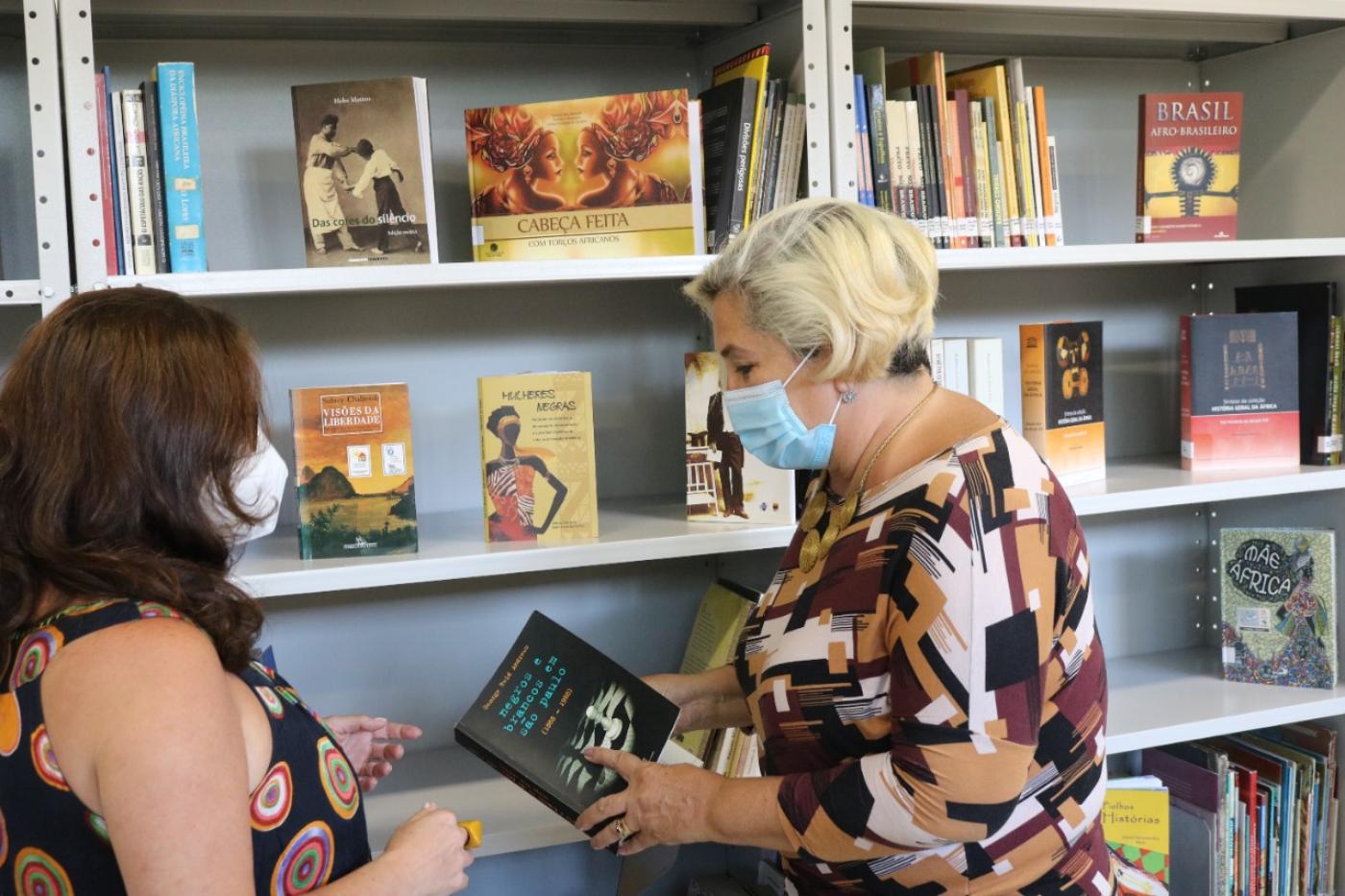 uma mulher , de máscara, em frente a outra mulher, que segura um livro #paratodosverem