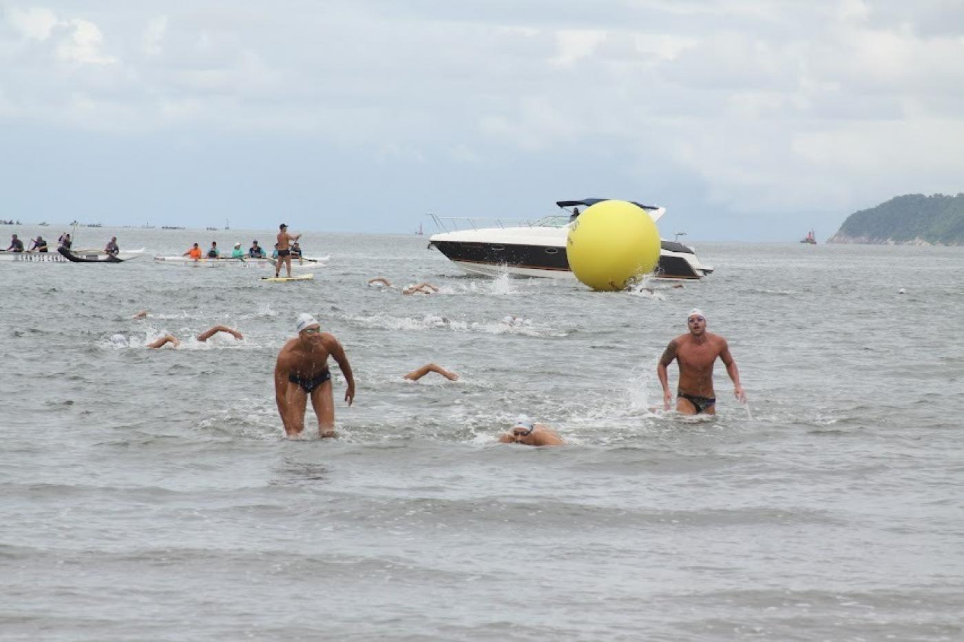 Trecho do mar com nadadores saindo da água, ao fundo uma lancha e uma grande boia amarela #paratodosverem