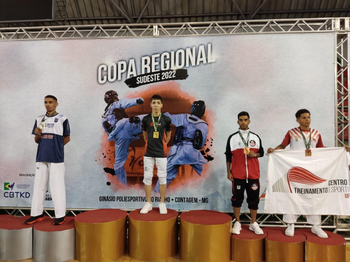 quatro atletas de pé em um pódio , o da direita tem uma bandeira aberta #paratodosverem