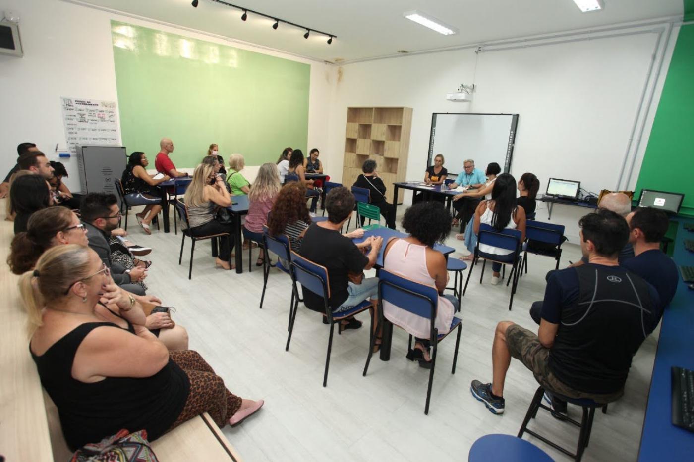 pessoas em uma sala sentadas em cadeiras escolares , ao fundo uma parede verde #paratodosverem