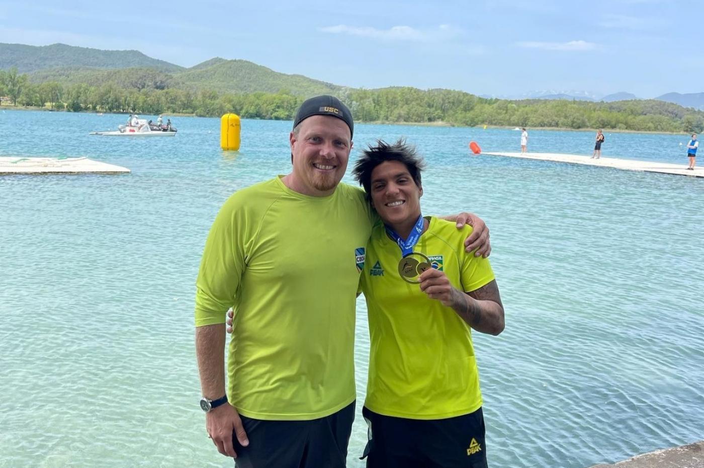treinador e ana marcela mostrando a medalha com mar ao fundo #paratodosverem