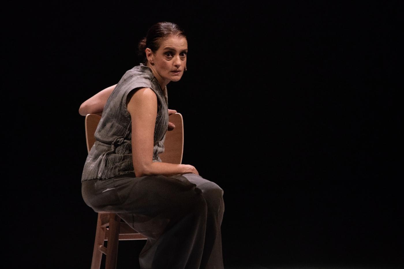 atriz sentada na cadeira olha pra frente com fundo escuro em palco #paratodosverem