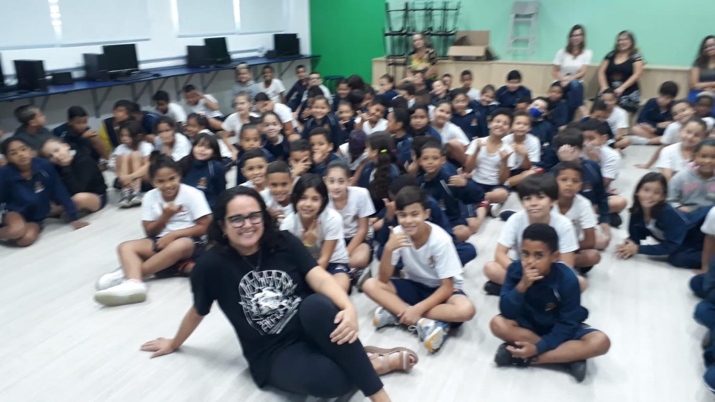 escritora e alunos estão sentados no chão posando para foto. #paratodosverem 