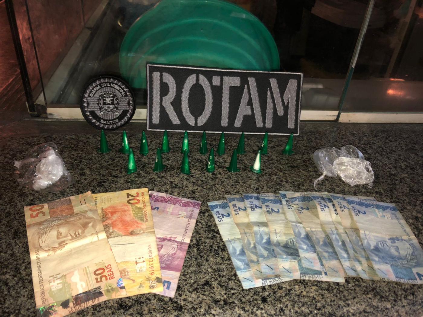 dinheiro e drogas sobre uma mesa com o distintivo da Rotam. #paratodosverem