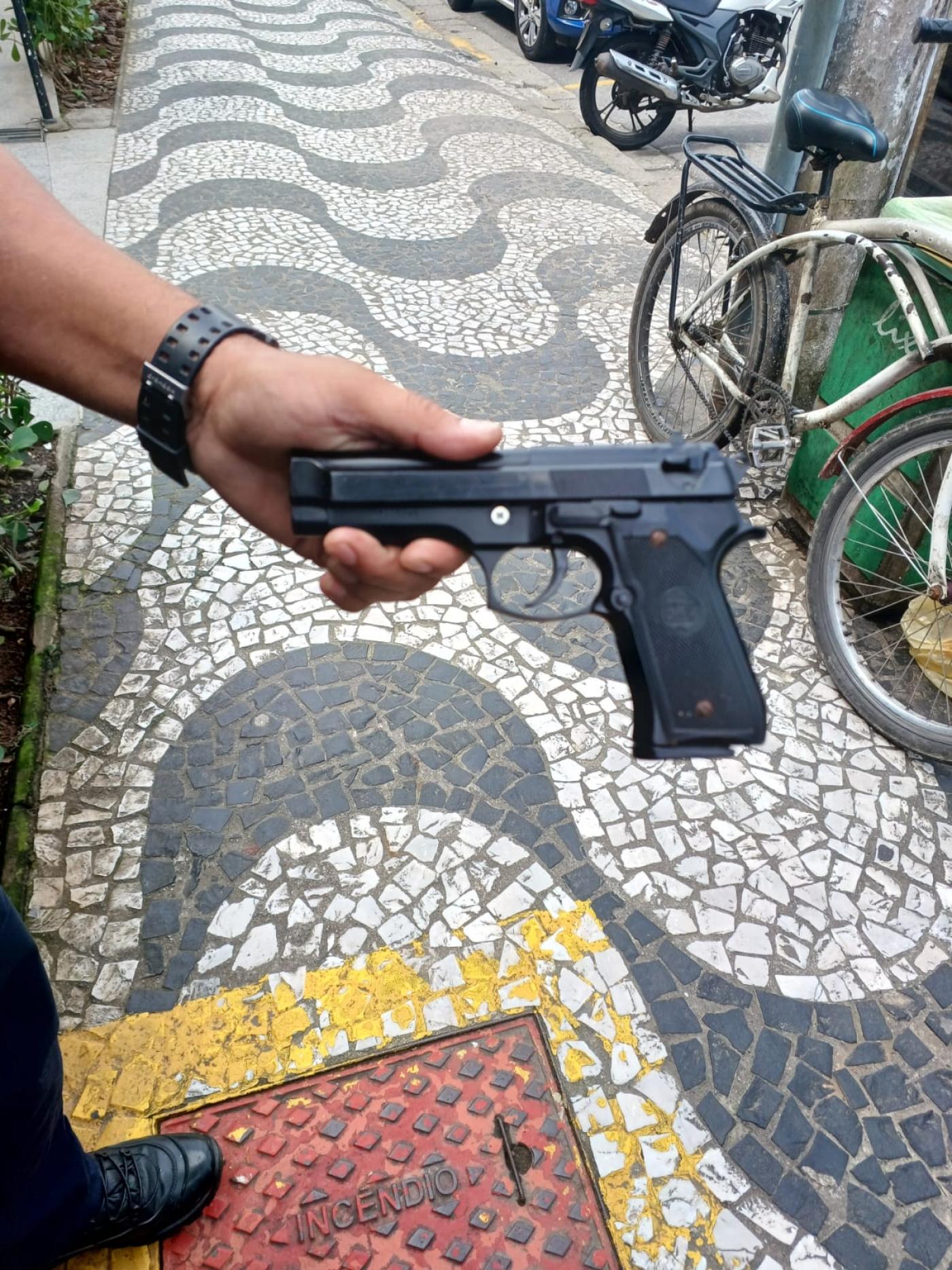 mão segura simulacro de arma. Há uma bicicleta encostada num poste ao fundo. #paratodosverem