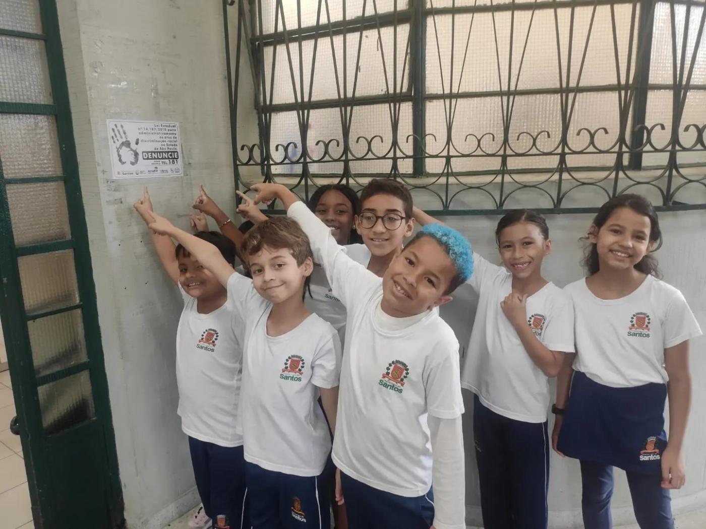 Sete crianças apontam ao mesmo tempo para cartaz afixado em parede. Elas sorriem para foto.#paratodosverem