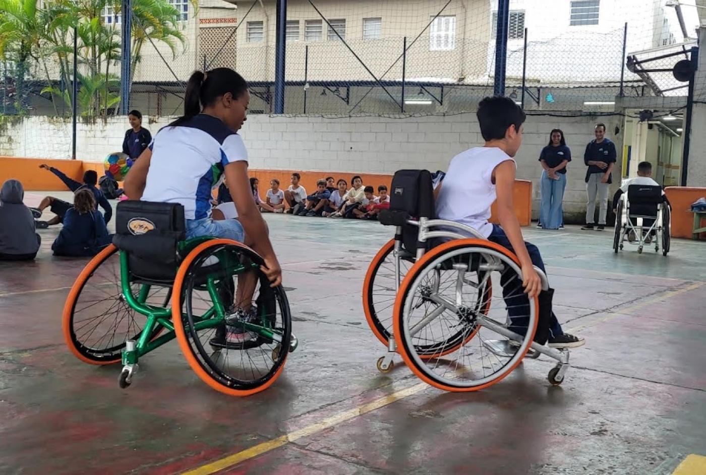 mulher e criança estão em cadeiras de rodas em quadra esportiva. #paratodosverem
