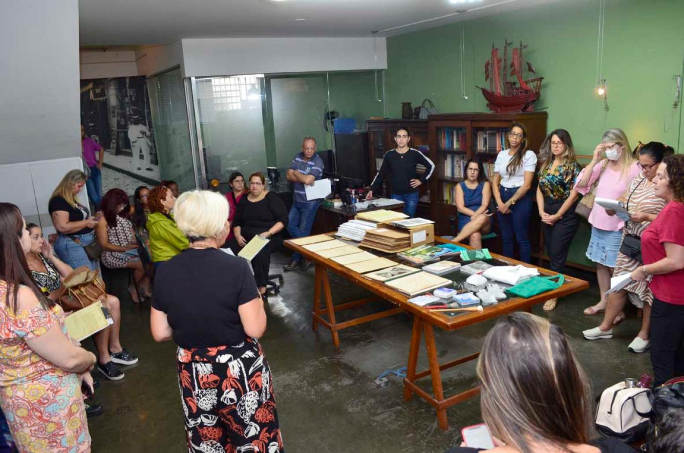 professores estão de pé em uma sala com uma grande mesa com documentos ao centro. #paratodosverem