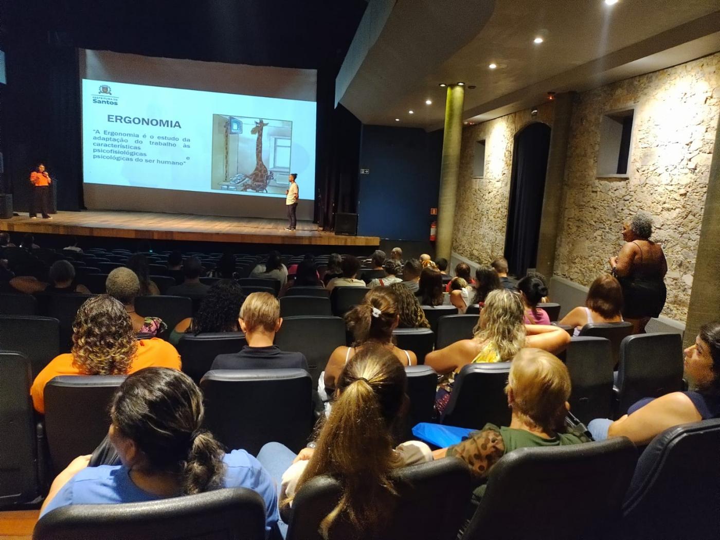várias pessoas sentadas em auditório assistindo palestra. Ao fundo, no palco, há um telão com imagem. #paratodosverem 